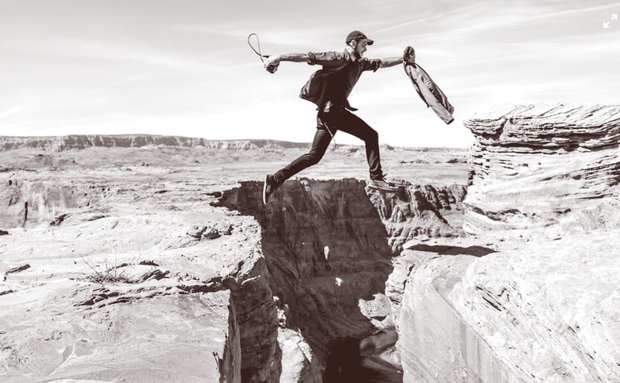 Black-and-white photograph of a man leaping across the gap between two rocky canyon cliffs