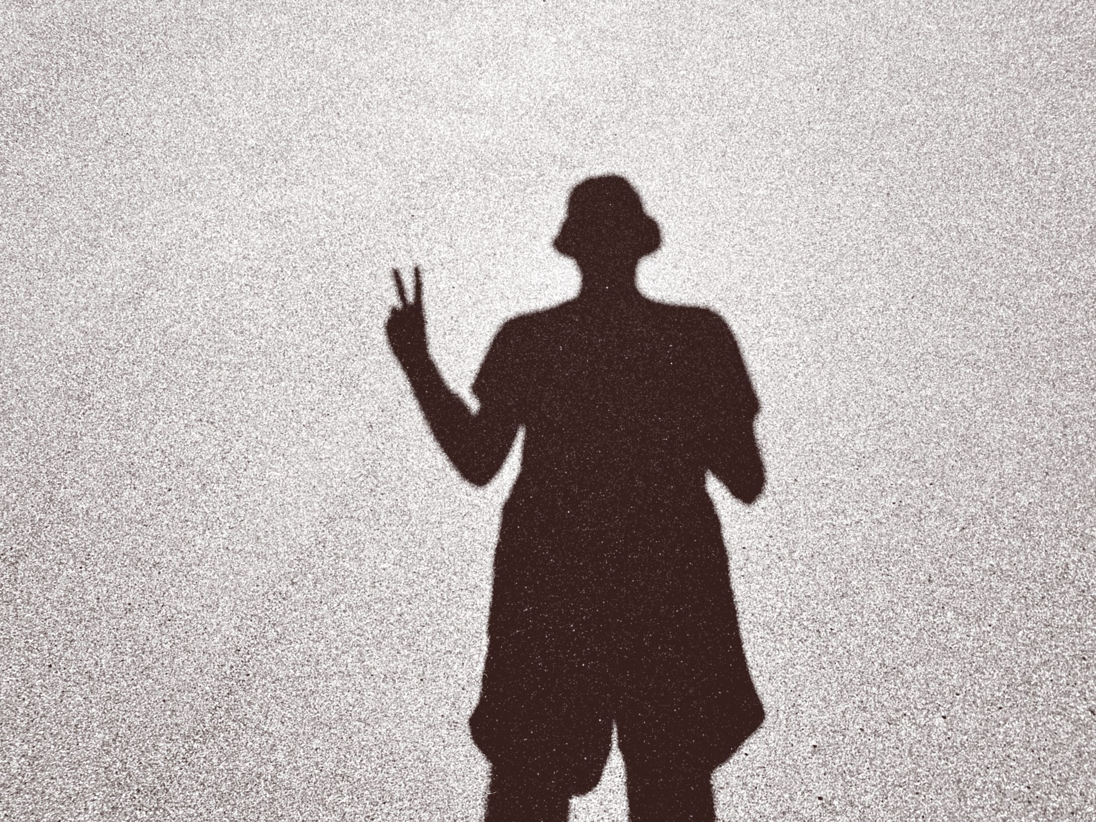 Black-and-white photograph of a person's shadow cast on textured ground, hand raised in a peace sign