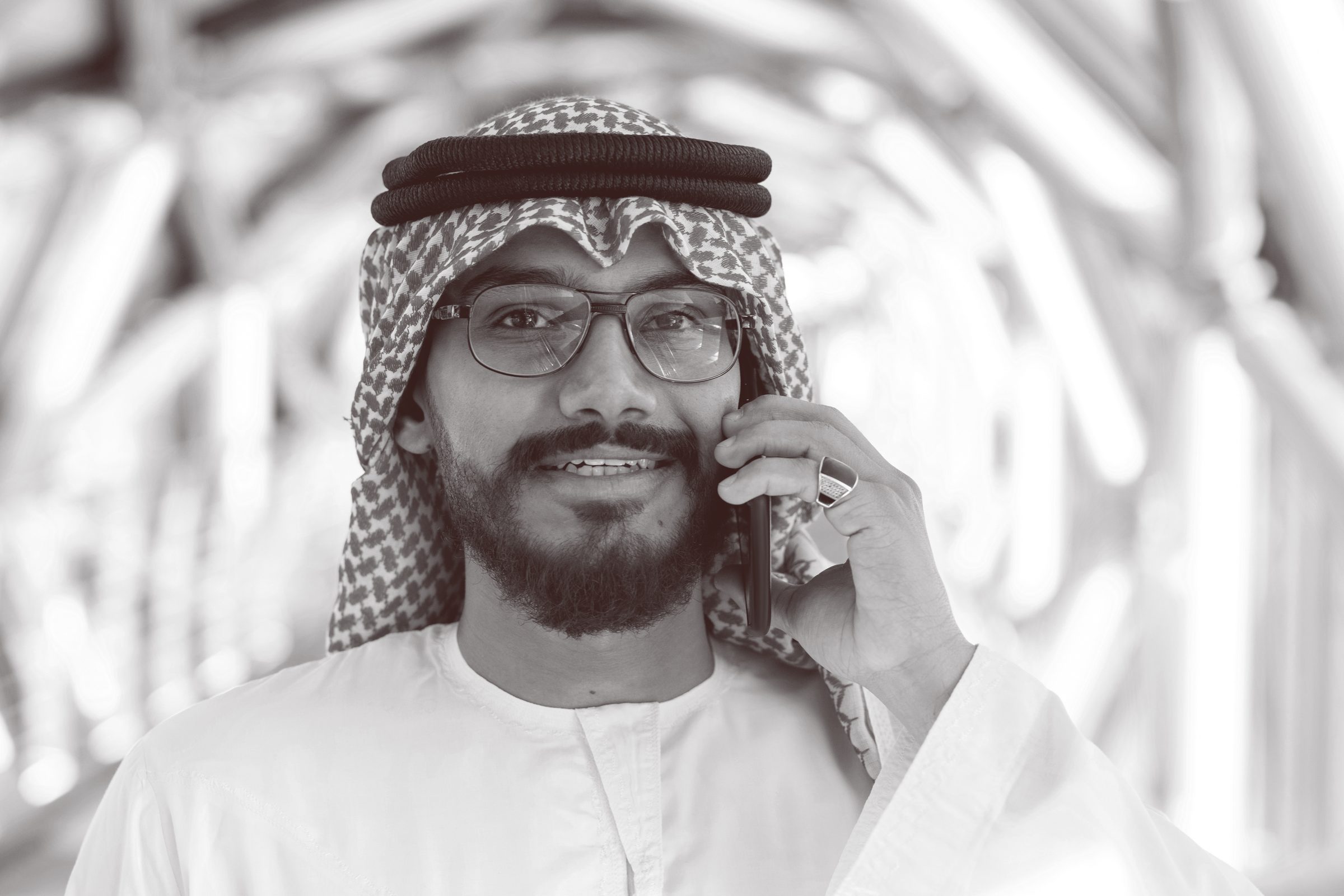Black-and-white portrait of a man in a ghutra and agal wearing glasses, speaking on his phone